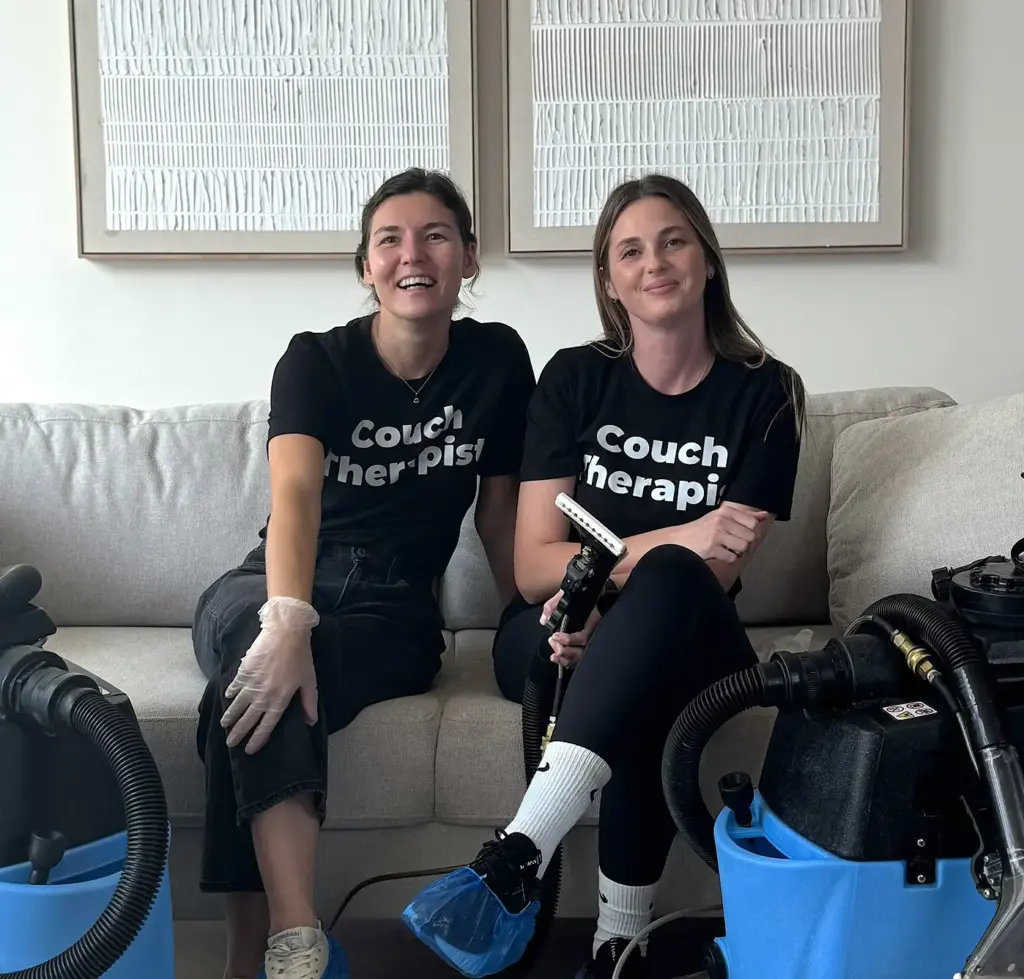Two smiling couch cleaning technicians sitting on a clean beige sofa next to their extraction machines