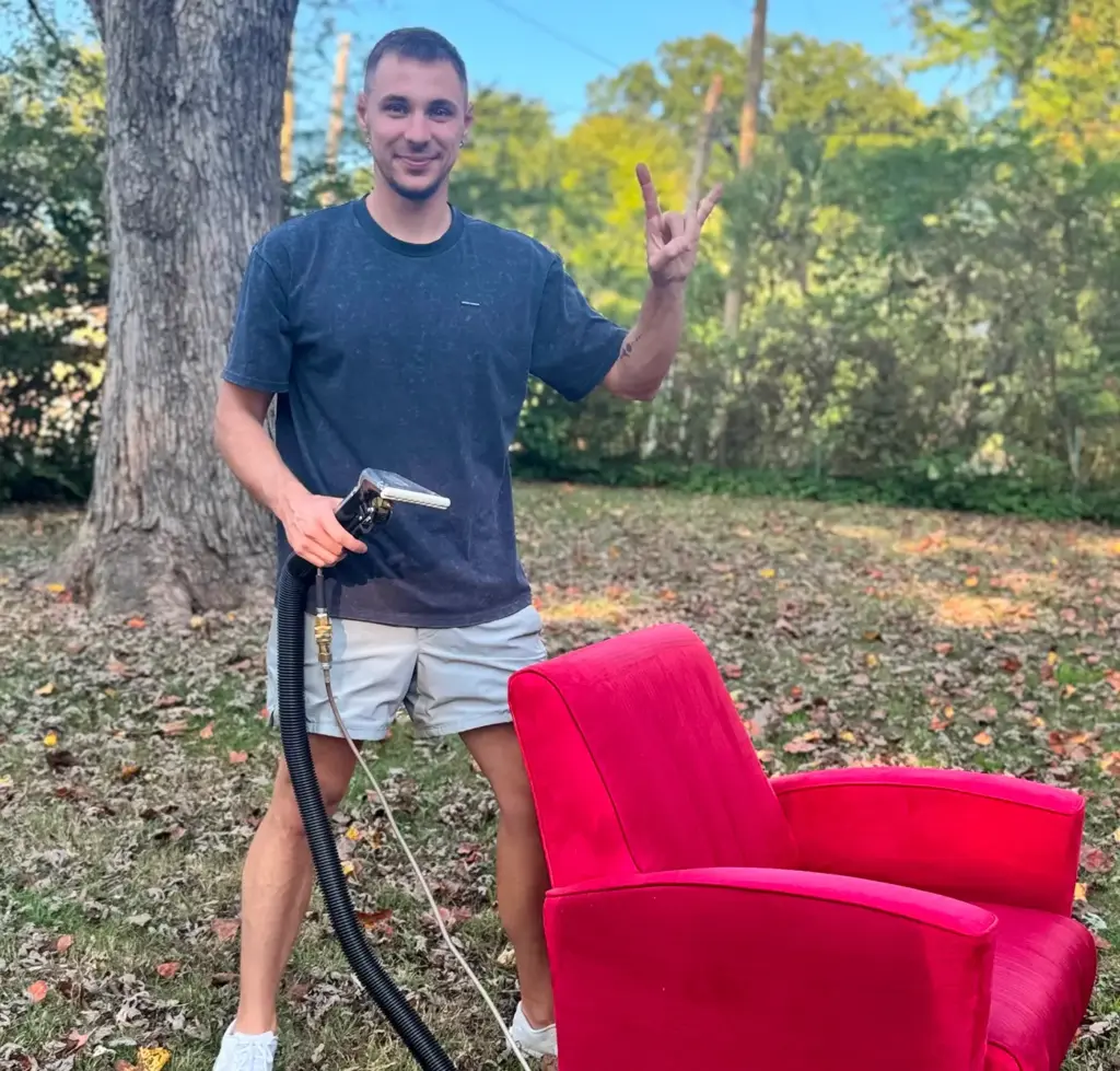 Smiling Texas Steamers technician holding a cleaning wand next to a freshly cleaned red armchair in Dallas, TX
